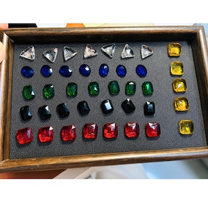 May include: A rectangular wooden display tray containing rows of faceted gemstones. The collection includes clear triangular stones, blue and green oval stones, black and red rectangular stones, and a vertical column of yellow square stones.