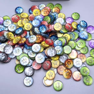Puede incluir: Una gran colección de fichas de juego circulares en varios colores, incluyendo azul, verde, amarillo, rojo y morado. Cada ficha presenta un número impreso, como 1 o 2, y un pequeño icono gráfico, esparcidas sobre una superficie clara.
