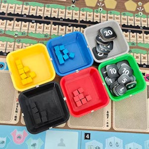 May include: Five square plastic trays in yellow, blue, red, black, and green, each containing small game components. The yellow, blue, red, and black trays hold small cubes, while the grey and green trays hold square tokens marked with numbers 5, 10, and 20.