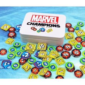May include: A white rectangular storage tin for the Marvel Champions card game, featuring the logo and game icons on the lid. The tin is surrounded by a collection of colorful acrylic game tokens, including yellow squares and circular red and blue markers.