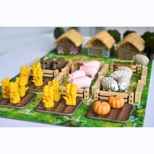 May include: A tabletop board game set featuring miniature farm elements including three small houses with thatched roofs, wooden fences, yellow wheat stalks, pink pigs, white sheep, and orange pumpkins arranged on a green field game board.