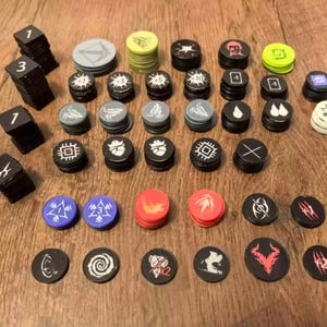 Puede incluir: Una colección de varios fichas y marcadores de juego, incluyendo bloques cuadrados negros con números y numerosos discos redondos en colores como negro, rojo, azul, verde y gris, con diversos símbolos e iconos gráficos en blanco.