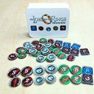 May include: A white storage box labelled The Lord of the Rings The Card Game with a collection of circular and square game tokens. The tokens feature various designs including numbers, glowing blue orbs, red patterns, and green landscape imagery.