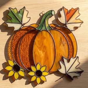 May include: A stained glass suncatcher featuring a central orange pumpkin with a green stem, surrounded by two yellow sunflowers and three maple leaves in shades of green, orange, and cream, all connected by dark metal solder lines.