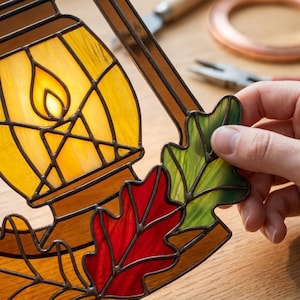 May include: A stained glass suncatcher featuring a yellow lantern with a flame design, surrounded by red and green oak leaves. The glass pieces are joined by dark metal solder lines, creating a decorative panel with autumn-inspired colors.