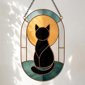 May include: Stained glass window art featuring a black cat silhouette. The cat is centered against a golden yellow circle, framed by teal, white, and clear glass. The piece is suspended by a chain.