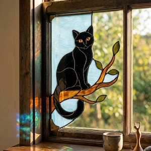 Könnte beinhalten: Ein Buntglas-Fensterbehang, der eine schwarze Katze mit orangefarbenen Augen zeigt, die auf einem braunen Ast mit grünen Blättern sitzt. Der Hintergrund besteht aus hellblauen Glasscheiben, eingefasst in dunkle Bleilinien aus Metall.