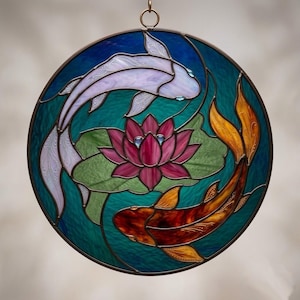 Pode incluir: Um apanhador de sol circular em vitral apresentando um peixe koi branco e um peixe koi laranja nadando em formação yin-yang ao redor de uma flor de lótus rosa central com vitórias-régias verdes sobre um fundo de água azul-petróleo.