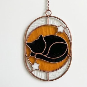 Op de afbeelding: Een ovaal glas-in-lood zonnevanger met een zwarte kat die zich tot een bolletje heeft opgerold tegen een ronde amberkleurige achtergrond. Het ontwerp bevat twee kleine witte sterren en is omlijst door een koperkleurige metalen rand.