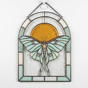May include: A stained glass window hanging featuring a pale mint green luna moth centred in front of a circular amber sun. The moth is framed by an arched border of white and mint glass panels, all held together by dark metal lead lines.