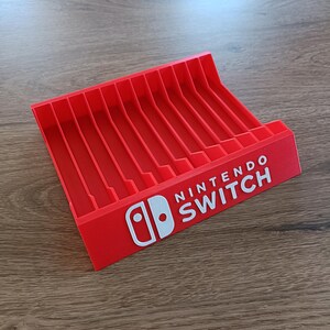 May include: A bright red plastic storage rack for game cartridges featuring ten vertical slots. The front panel displays the white Nintendo Switch logo and text. The structure is angled to hold items securely in a row, with a smooth, matte finish.