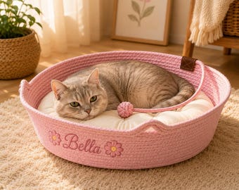 Puede incluir: Una cama redonda para mascotas hecha de cuerda trenzada rosa con un cojín acolchado de color crema en su interior. El exterior de la cama está bordado con el nombre Bella en letra cursiva, flanqueado por dos pequeñas flores rosas y amarillas.