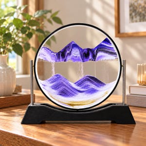 Puede incluir: Un expositor de arte en arena circular con capas de arena morada, blanca y dorada dentro de un marco de cristal transparente. El marco se apoya en una base negra con dos varillas metálicas plateadas, creando un efecto de paisaje montañoso.
