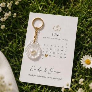 May include: A white rectangular card featuring a June calendar with a gold heart on the 24th, the names Emily & Simon, and a gold keychain with a clear shell charm containing a white pearl attached to the top left corner of the paper card.