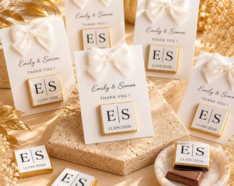 May include: Square chocolate favors wrapped in white paper with a gold border, featuring the letters ES and the date 11/09/2026. Each is attached to a white card with a white satin bow, printed with Emily & Simon and the text THANK YOU!