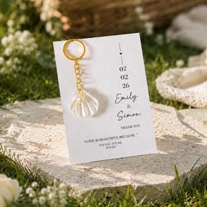Può includere: Un cartoncino rettangolare bianco con un portachiavi dorato e un ciondolo a forma di conchiglia bianca. Il cartoncino riporta la data 07 02 26, i nomi Emily & Simon e il testo: THANK YOU. LOVE IS BEAUTIFUL BECAUSE... IT'S YOU, IT'S ME, IT'S US.
