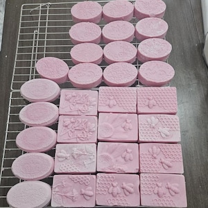 May include: Pink handmade soaps in oval and rectangular shapes, arranged on a wire rack. The soaps feature embossed designs of flowers, bees, and honeycomb patterns. The soaps are a soft pink color.