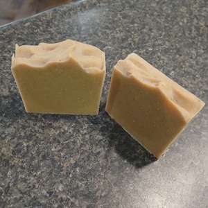 May include: Two bars of handmade soap. The soap is a light tan color with a textured top. The bars are rectangular with a slightly wavy top edge. The soap is sitting on a dark gray speckled surface.