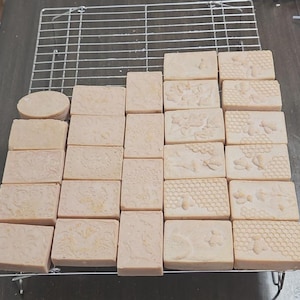 May include: A collection of handmade soaps in various shapes and sizes, resting on a metal cooling rack. The soaps are light beige with embossed designs, including floral patterns, bees, and honeycomb textures. One soap is round.