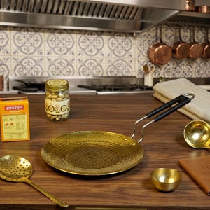 May include: A round, hammered brass tawa pan with a long metal handle featuring black grips, accompanied by a brass slotted spoon, a brass ladle, a small brass bowl, a yellow spice box with text, and a glass jar filled with nuts.