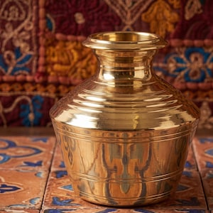 May include: A polished brass pot with a wide, rounded body and a narrow neck that flares into a circular rim. The surface features horizontal engraved lines around the midsection and neck, creating a smooth, reflective metallic finish.