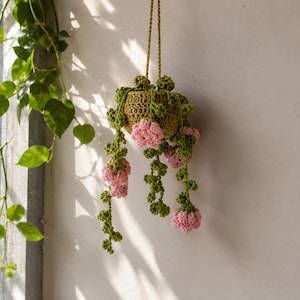 May include: A crocheted hanging plant in a textured olive green pot. The plant features long, trailing green vines with small, rounded leaves and clusters of soft pink flowers. The entire piece is suspended by a thin, matching olive green cord.