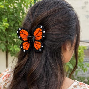 May include: A butterfly-shaped hair claw clip featuring a black and orange crocheted design. The wings are outlined in black with orange interior sections and small white beads attached along the outer edges of the butterfly wings.