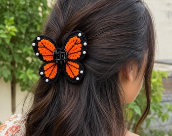 Könnte beinhalten: Eine schmetterlingsförmige Haarklammer mit einem gehäkelten Design in Schwarz und Orange. Die Flügel sind schwarz umrandet mit orangefarbenen Innenbereichen und kleinen weißen Perlen, die entlang der Außenkanten der Schmetterlingsflügel befestigt sind.