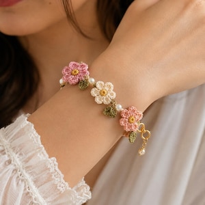 以下が含まれることがあります： ピンクとクリーム色の花、黄色の中心、緑の葉、小さな真珠のアクセントが特徴の、かぎ針編みのブレスレット。ブレスレットは、金色の留め金とぶら下がっている真珠のチャームで固定されています。