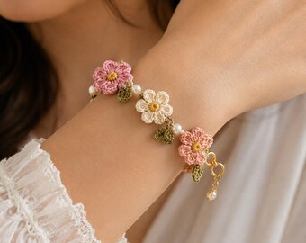 Könnte beinhalten: Ein gehäkeltes Armband mit rosa und cremefarbenen Blumen mit gelben Zentren, grünen Blättern und kleinen Perlenakzenten. Das Armband wird mit einem goldfarbenen Verschluss und einem baumelnden Perlenanhänger befestigt.