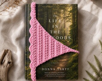 Könnte beinhalten: Ein handgefertigter, rosa Häkel-Lesezeichen liegt auf einem Buch mit dem Titel "The Life in the Woods" von Donna Tartt. Das Lesezeichen hat eine dreieckige Form mit einem gewellten Rand. Das Buchcover zeigt eine Waldszene.