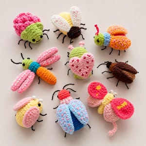 May include: A collection of nine small, handcrafted crochet insects and bugs in various bright colours including pink, yellow, green, blue, and brown. Each figure features detailed stitching, small black eyes, and thin dark antennae or legs.