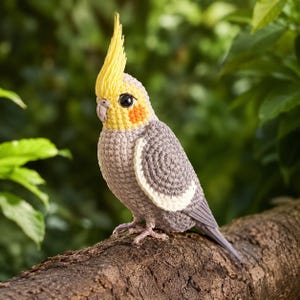 Puede incluir: Una figura de cacatúa ninfa tejida a ganchillo con cuerpo gris, cresta amarilla, cara amarilla con manchas naranjas en las mejillas y una franja blanca en el ala. El pájaro tiene un pequeño pico gris y ojos negros, hechos con puntos visibles.