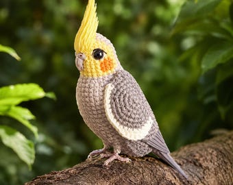 Könnte beinhalten: Eine gehäkelte Nymphensittich-Figur mit grauem Körper, gelber Haube, gelbem Gesicht mit orangefarbenen Wangenflecken und einem weißen Flügelstreifen. Der Vogel hat einen kleinen grauen Schnabel und schwarze Augen, gefertigt mit sichtbaren Maschen.