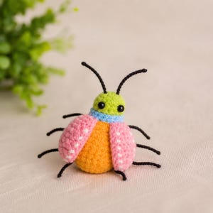 May include: A small, handcrafted crochet beetle toy featuring a bright lime green head, a light blue neck band, a solid orange body, and two pink wings decorated with small white dots. The insect has six thin black legs and two black antennae.