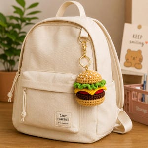 May include: A cream-colored backpack with a crocheted hamburger keychain. The keychain features a detailed miniature burger with a sesame seed bun, lettuce, tomato, and a beef patty. The backpack has a front pocket with the text "DAILY PRACTICE WITH SUNSHINE".