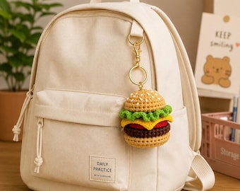 Könnte beinhalten: Ein cremefarbener Rucksack mit einem gehäkelten Hamburger-Schlüsselanhänger. Der Schlüsselanhänger zeigt einen detaillierten Miniatur-Burger mit Sesambrötchen, Salat, Tomate und einem Rindfleisch-Patty. Der Rucksack hat eine Vordertasche mit dem Text "DAILY PRACTICE WITH SUNSHINE".