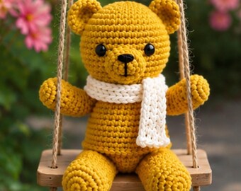Könnte beinhalten: Ein handgehäkelter Teddybär in Senfgelb mit einem weißen Schal, der auf einer Holzschaukel sitzt. Der Bär hat schwarze Augen und eine schwarze Nase. Die Schaukel ist an einem braunen Seil aufgehängt.