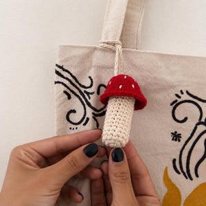 May include: A beige canvas tote bag featuring black swirling line art and a yellow graphic element. Attached to the bag handle is a small, three-dimensional crocheted mushroom charm with a red cap featuring white dots and a cream-colored stem.