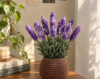 Könnte beinhalten: Eine gehäkelte Lavendelpflanze mit mehreren violetten Blütenähren und grünen Stielen, eingetopft in einen runden, strukturierten braunen Häkeltopf. Die Pflanze besteht aus verschiedenen Violetttönen und wirkt wie eine handgefertigte Blume.