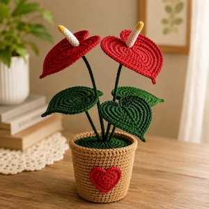 May include: A crocheted plant with two red anthurium flowers, green leaves, and a tan pot with a red heart. The flowers have white and yellow details. The plant is on a wooden surface.