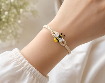 May include: A cream-colored macrame bracelet featuring a yellow, black, and white bee charm. The bee charm is centered on the bracelet. The bracelet is worn on a wrist.