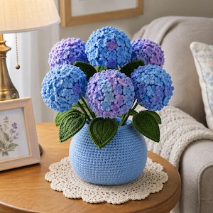 May include: A crochet hydrangea arrangement featuring multiple spherical flower heads in shades of blue and purple, accompanied by dark green crocheted leaves. The flowers are displayed in a round, light blue crocheted vase sitting on a white doily.