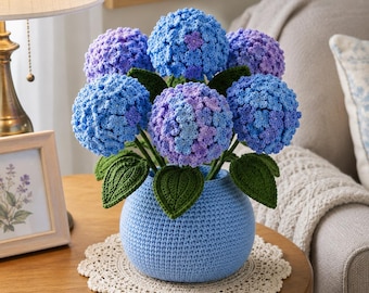 Könnte beinhalten: Ein gehäkeltes Hortensien-Arrangement mit mehreren kugelförmigen Blütenköpfen in Blau- und Violetttönen, begleitet von dunkelgrünen gehäkelten Blättern. Die Blumen stehen in einer runden, hellblauen gehäkelten Vase auf einer weißen Spitze.