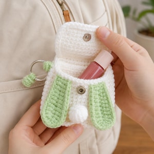 Puede incluir: Una pequeña bolsa de ganchillo blanca con cierre de solapa, que presenta dos orejas colgantes de color verde claro y una pequeña cola de pompón blanca. La bolsa incluye un llavero de metal plateado y un botón a presión, con un frasco rosa dentro.