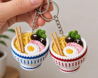 Könnte beinhalten: Zwei gehäkelte Ramen-Schüssel-Schlüsselanhänger mit gelben Nudeln, einem weißen Ei mit gelbem Kern, rosa Fischkuchenstücken, grünem Brokkoli und braunen Pilzen. Eine Schüssel hat blaue Streifen, die andere rote, beide enthalten hölzerne Essstäbchen.