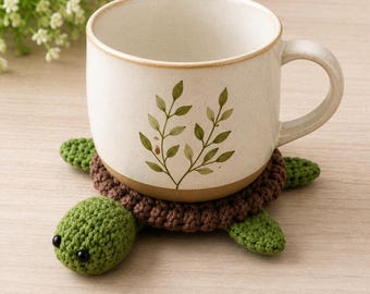 Könnte beinhalten: Eine cremefarbene Keramiktasse mit braunem Boden und grünem Blattmuster, die auf einem handgefertigten Schildkröten-Untersetzer aus Häkelarbeit steht. Der Untersetzer hat einen braunen Panzer und einen grünen Kopf und Flossen. Die Tasse hat einen gebogenen Griff.