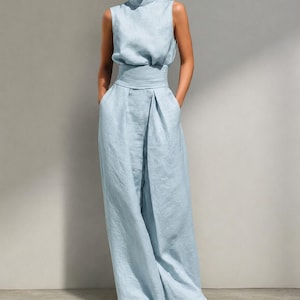 May include: A pale blue sleeveless jumpsuit with a high neckline and wide-leg trousers. The outfit has a cinched waist and pockets. The model is wearing black sandals. The fabric appears to be linen or a similar lightweight material.