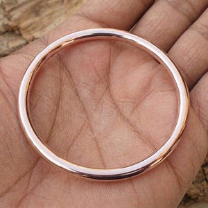 以下が含まれることがあります： ローズゴールドカラーのバングルブレスレット。円形のブレスレットは滑らかで光沢があり、一貫した色合いです。バングルは手で持たれており、背景には木製の表面があります。このシンプルでエレガントなブレスレットは、日常使いに適しています。