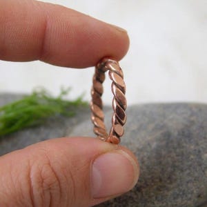 Pode incluir: Um anel cor de cobre com um design de corda torcida. O anel é segurado entre os dedos, mostrando seu intrincado padrão trançado. O tom quente do anel contrasta com o fundo neutro, destacando o artesanato da joia.